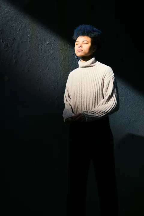 A woman in a cream turtleneck sweater standing near a sunlit wall.