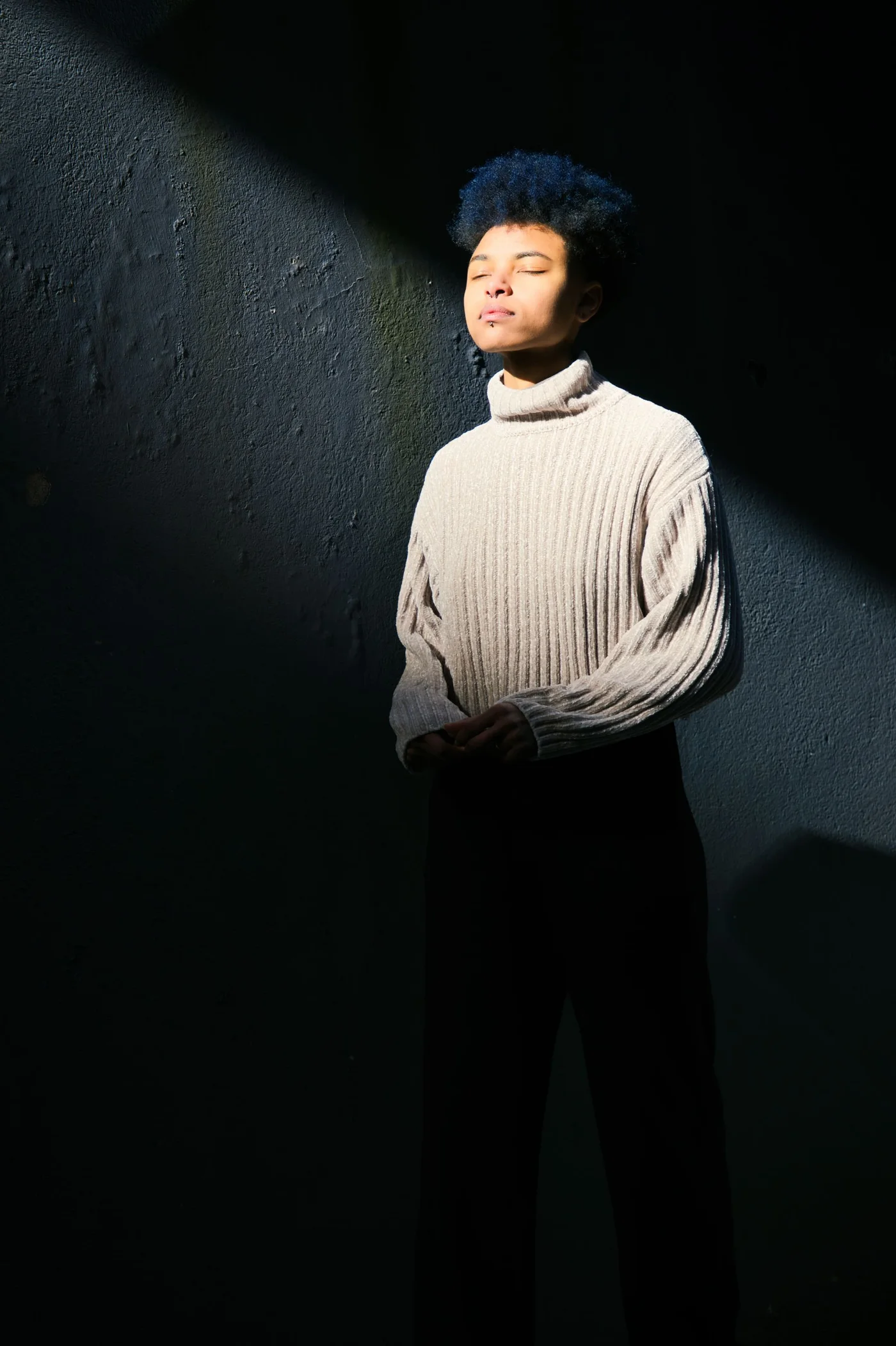 A woman in a cream turtleneck sweater standing near a sunlit wall.
