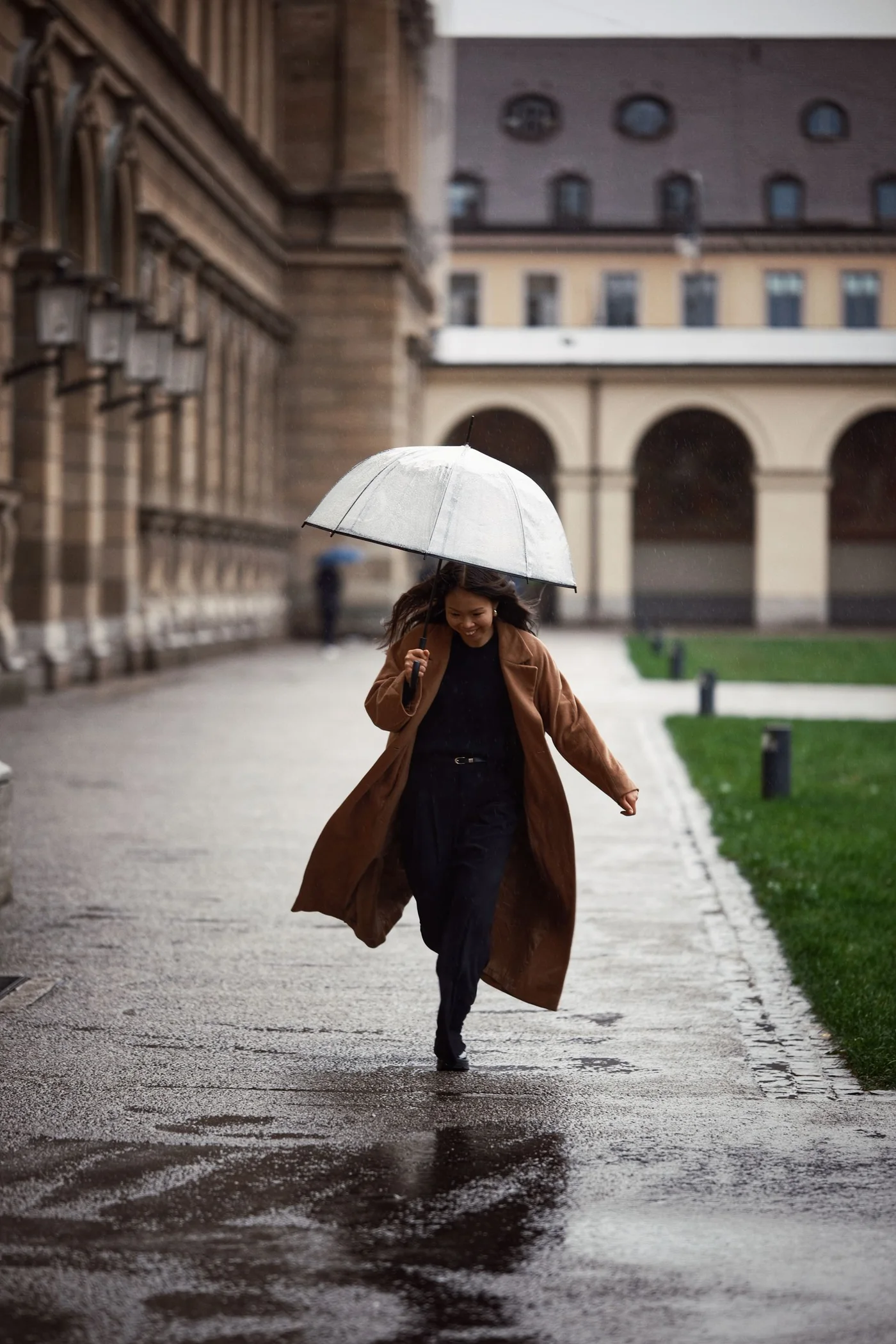 A woman in a brown coat walking with an umbrella along a wet city street.