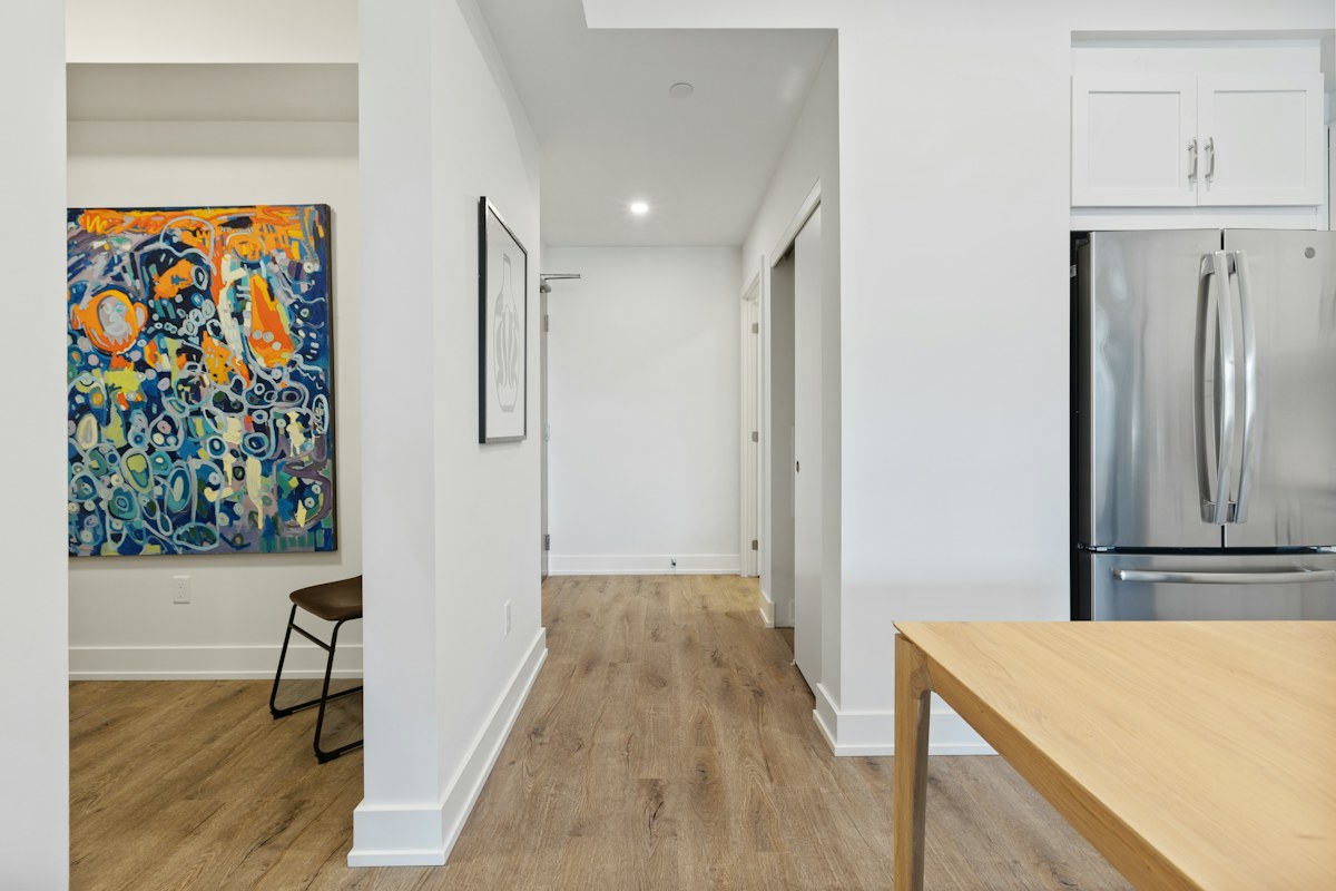 A pale hallway adjoining a kitchen, with timber flooring and a colourful framed picture.
