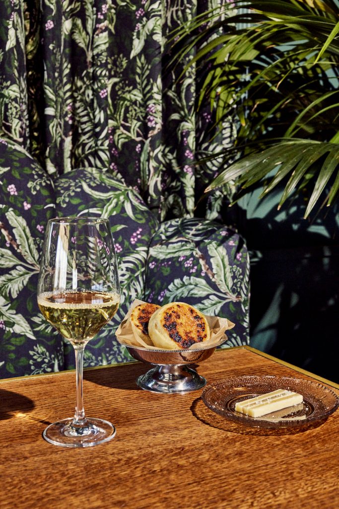 Glass of white wine, silver dish with pastries, and cheese on wood-topped table in front of botanical wallpaper.