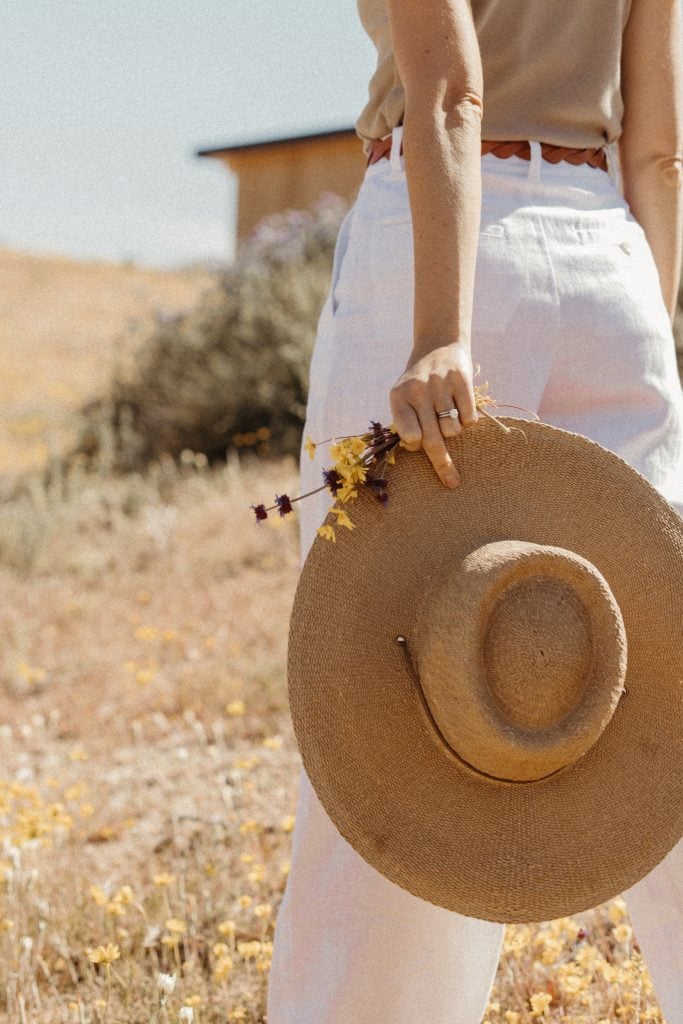 Brunette woman wearing white linen pants carrying sunhat in field.